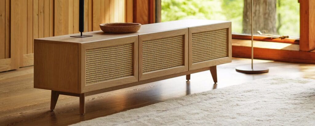 A modern wooden media cabinet with cane front panels, set on four tapered legs, placed near a large window with outdoor greenery visible.